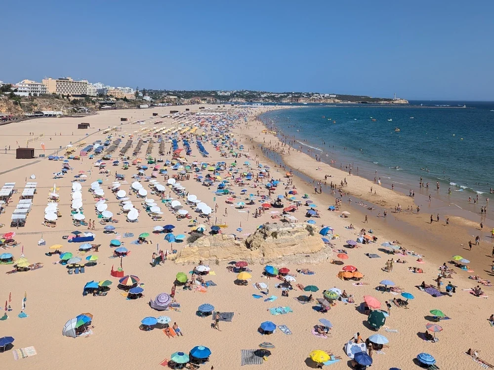 An aerial view of a bustling, sun-drenched beach in Praia da Rocha, packed with colorful umbrellas and beachgoers enjoying the coast.