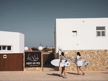 Two surfers carrying their boards walk towards the entrance of the stylish Sagres Sun Stay hostel accommodation.