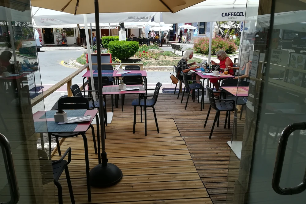 Guests dining on the sunny outdoor patio of Scoopit Food Atelier restaurant in Tavira, with a wooden deck and umbrellas.