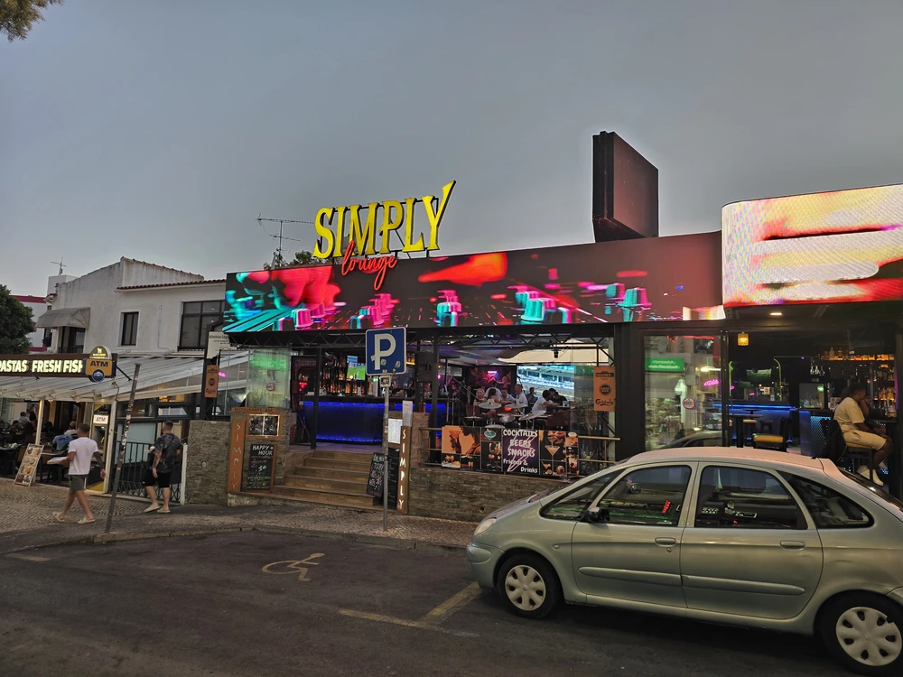The vibrant exterior of Simply Bar on the main strip in Albufeira at dusk, with colorful neon signs lighting up the bustling nightlife scene.