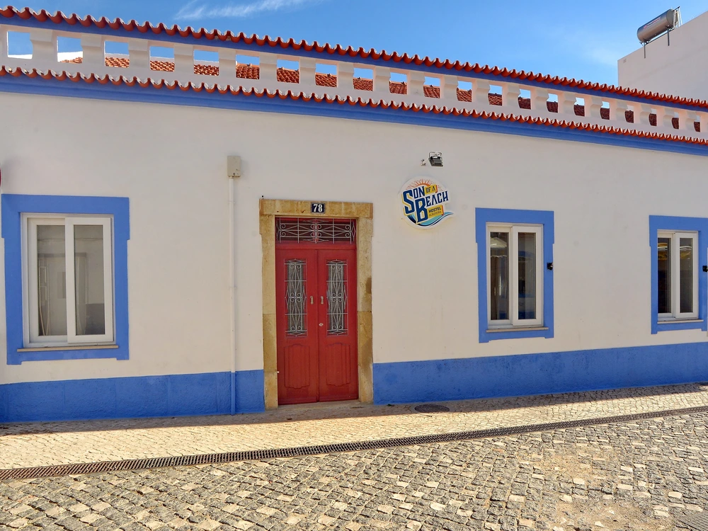 The charming white and blue facade of the Son of a Beach hostel in Albufeira, featuring a traditional red door on a classic cobblestone street.
