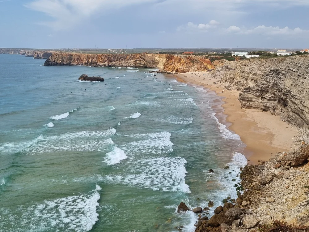 Surfers enjoying the waves on a scenic beach in Sagres, with dramatic golden cliffs lining the shore.