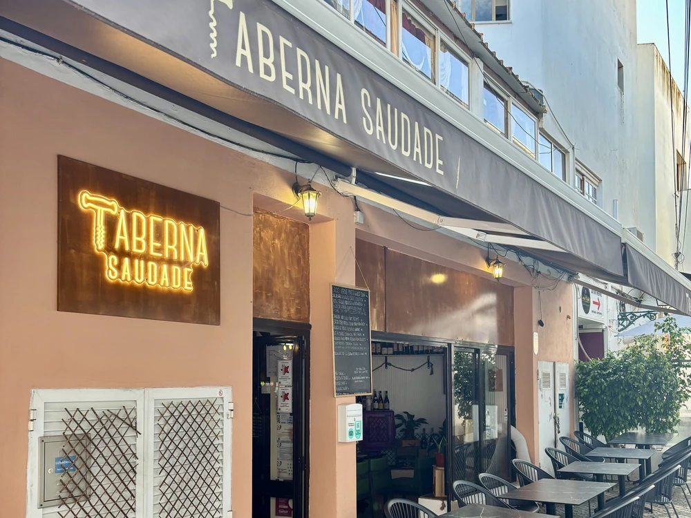 The stylish entrance to Taberna Saudade, a modern restaurant in Albufeira, featuring a warm, illuminated sign and an inviting outdoor dining area.