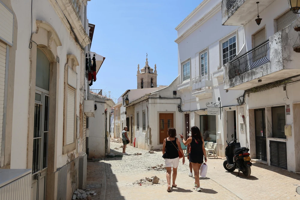 Visitors walking down a narrow, charming cobblestone alley in the historic Tavira city streets, with a church tower visible in the distance.