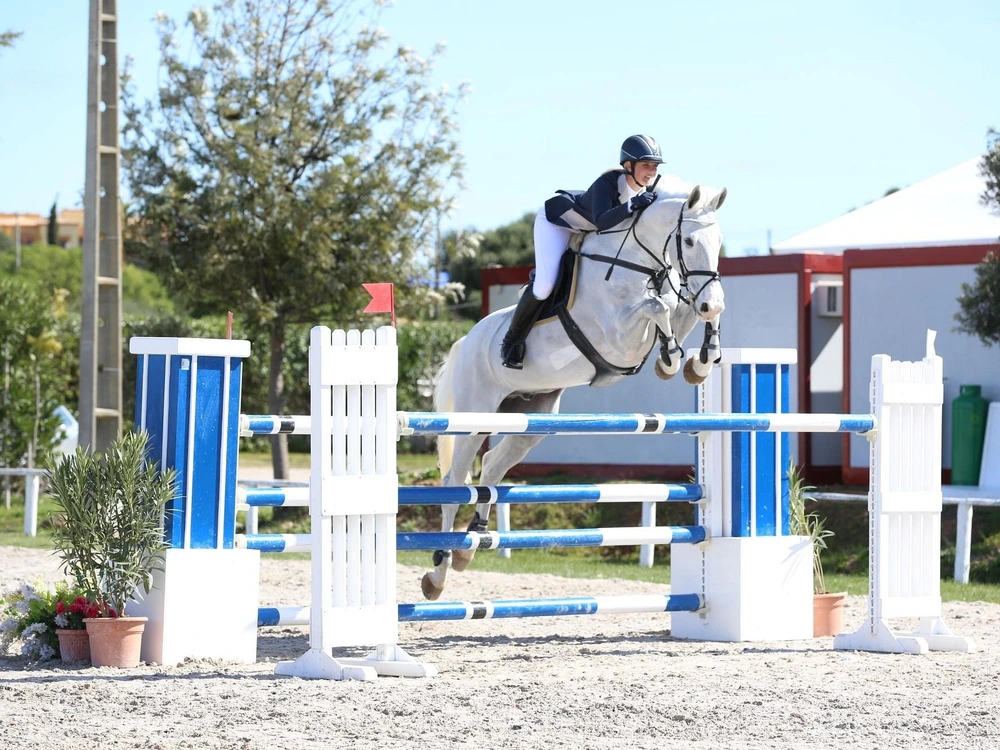A skilled rider on a white horse clearing a jump during the Vilamoura Champions Tour at the Vilamoura Equestrian Centre.