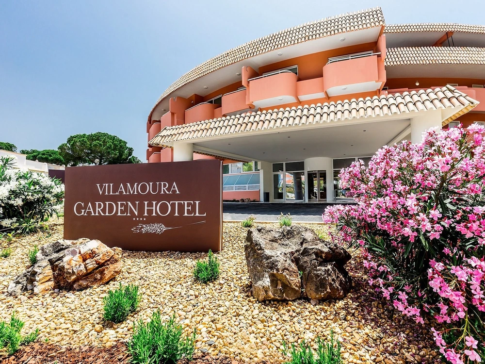 The welcoming entrance to the Vilamoura Garden Hotel, showing the main sign surrounded by landscaped gardens and pink flowers.